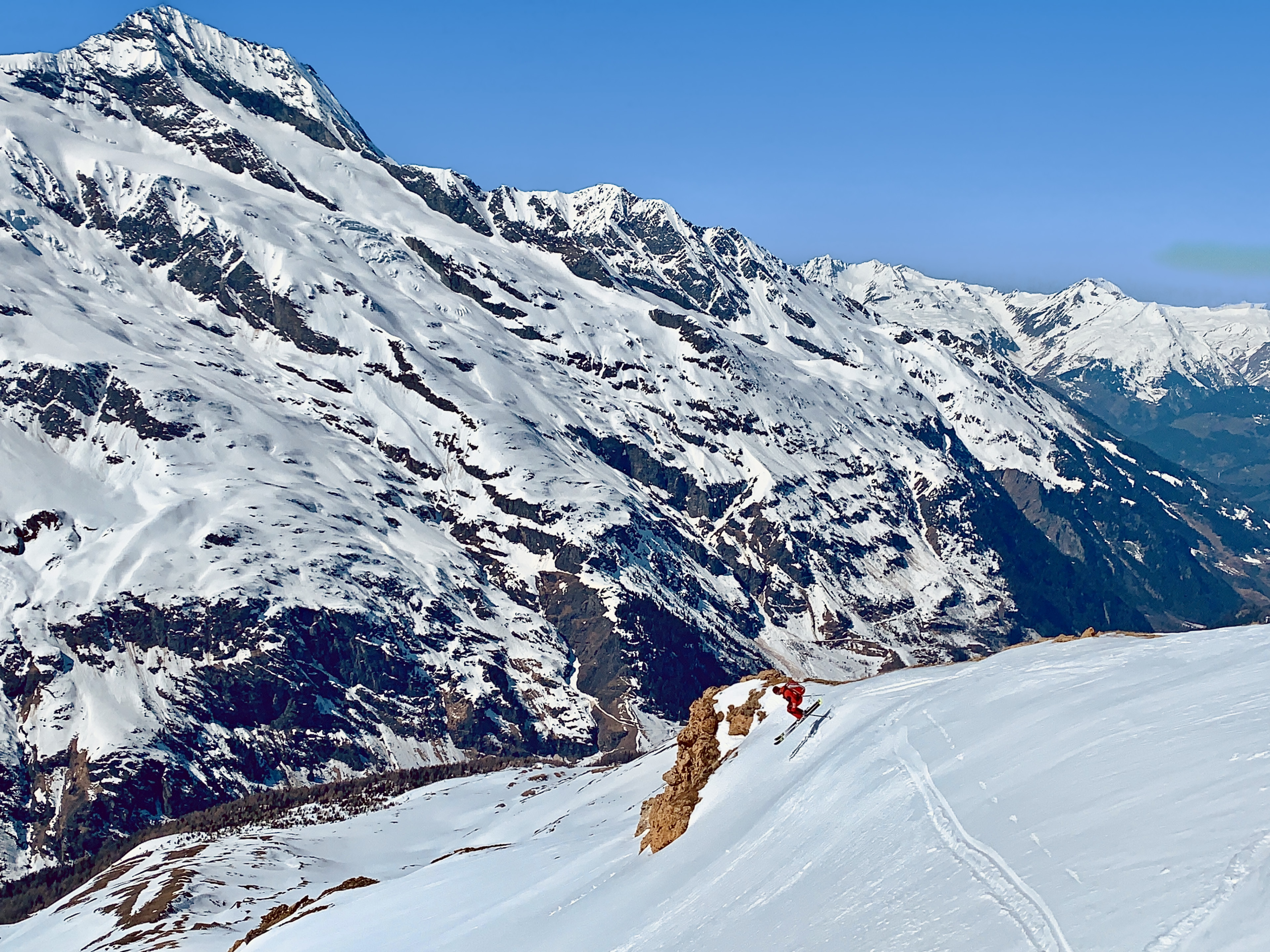 Heliskiing im Hochgebirge: Skifahrer im roten Anzug springt von einer Felskante, vor verschneitem Berg und langem Tal