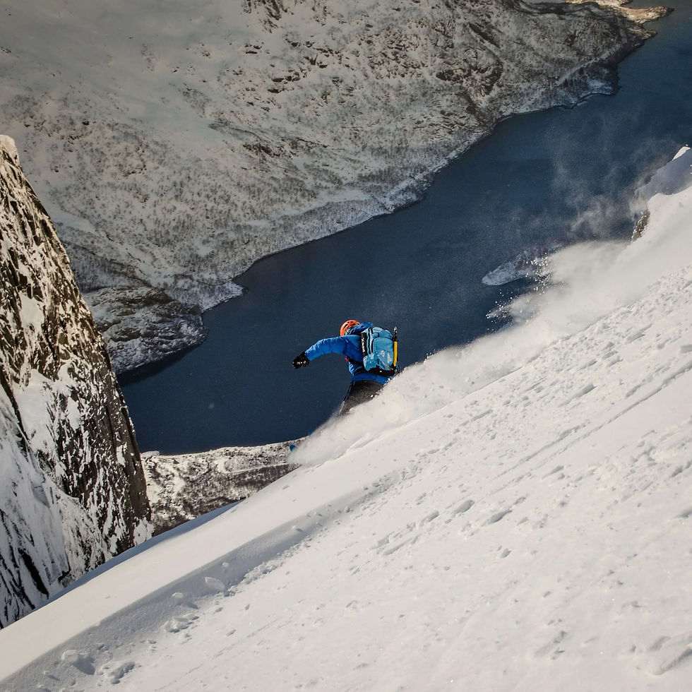 A skier in an orange helmet mid-turn on a steep slope, a plume of snow behind him; below his skis the slope drops straight to the dark fjord below, a vertical rock wall to the left