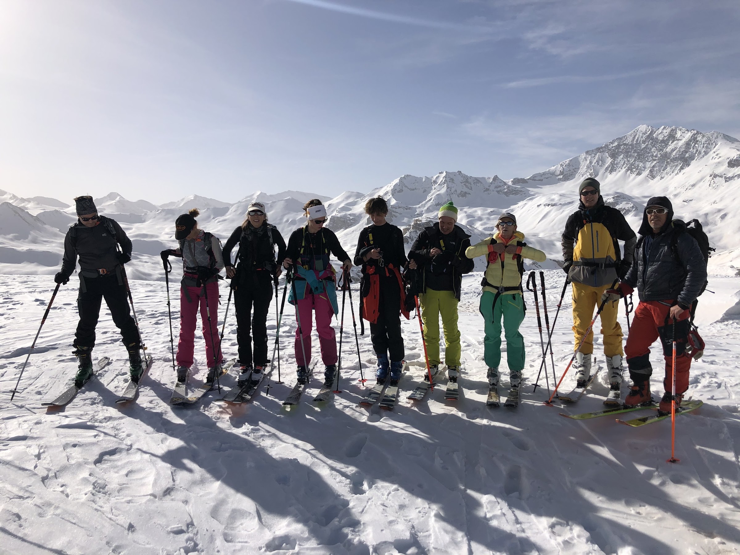 Neun Skitourengeher stellen sich für ein Gruppenfoto auf, dahinter verschneite Gipfel