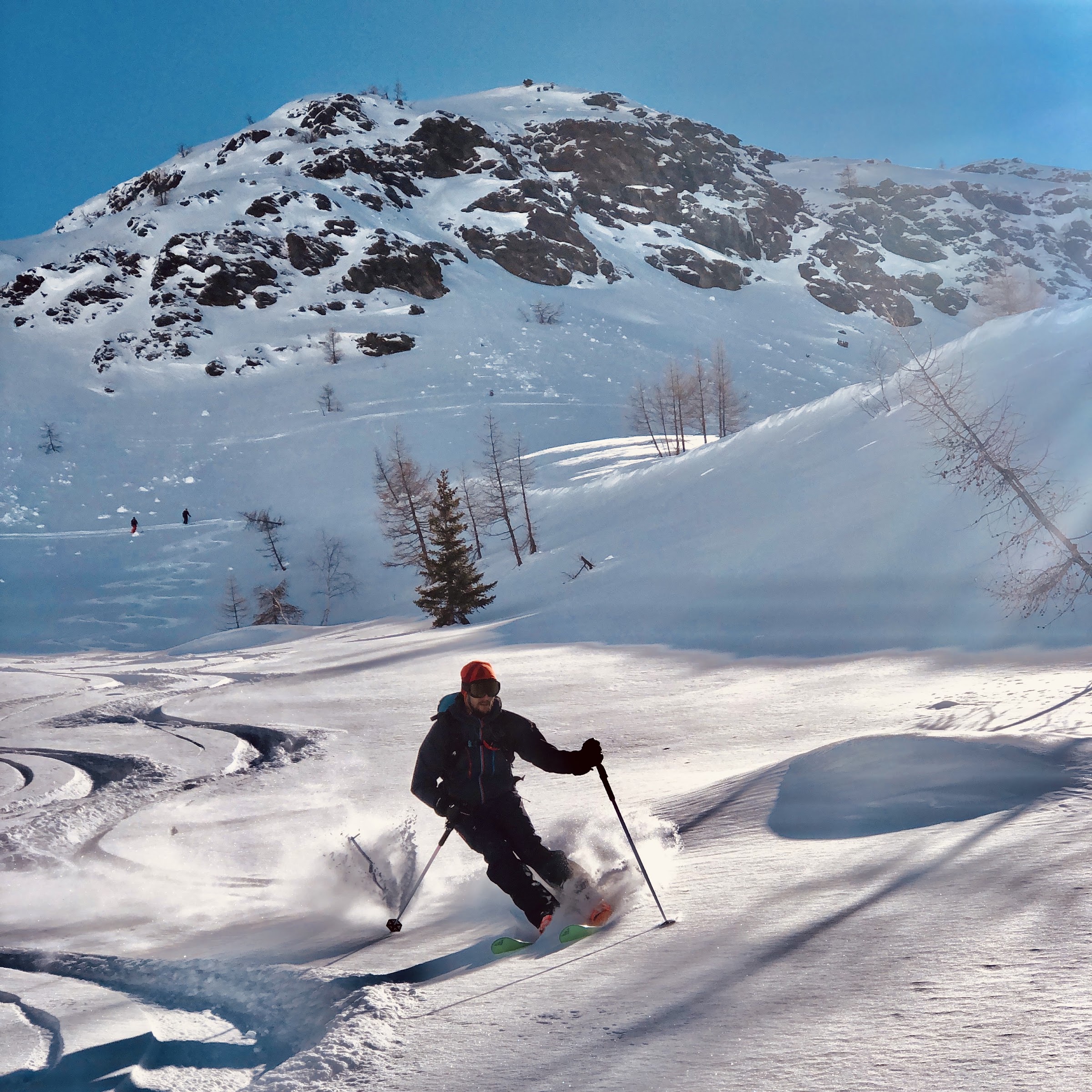 Skifahrer im Schwung durch Pulverschnee unter einem felsigen Hang mit Lärchen