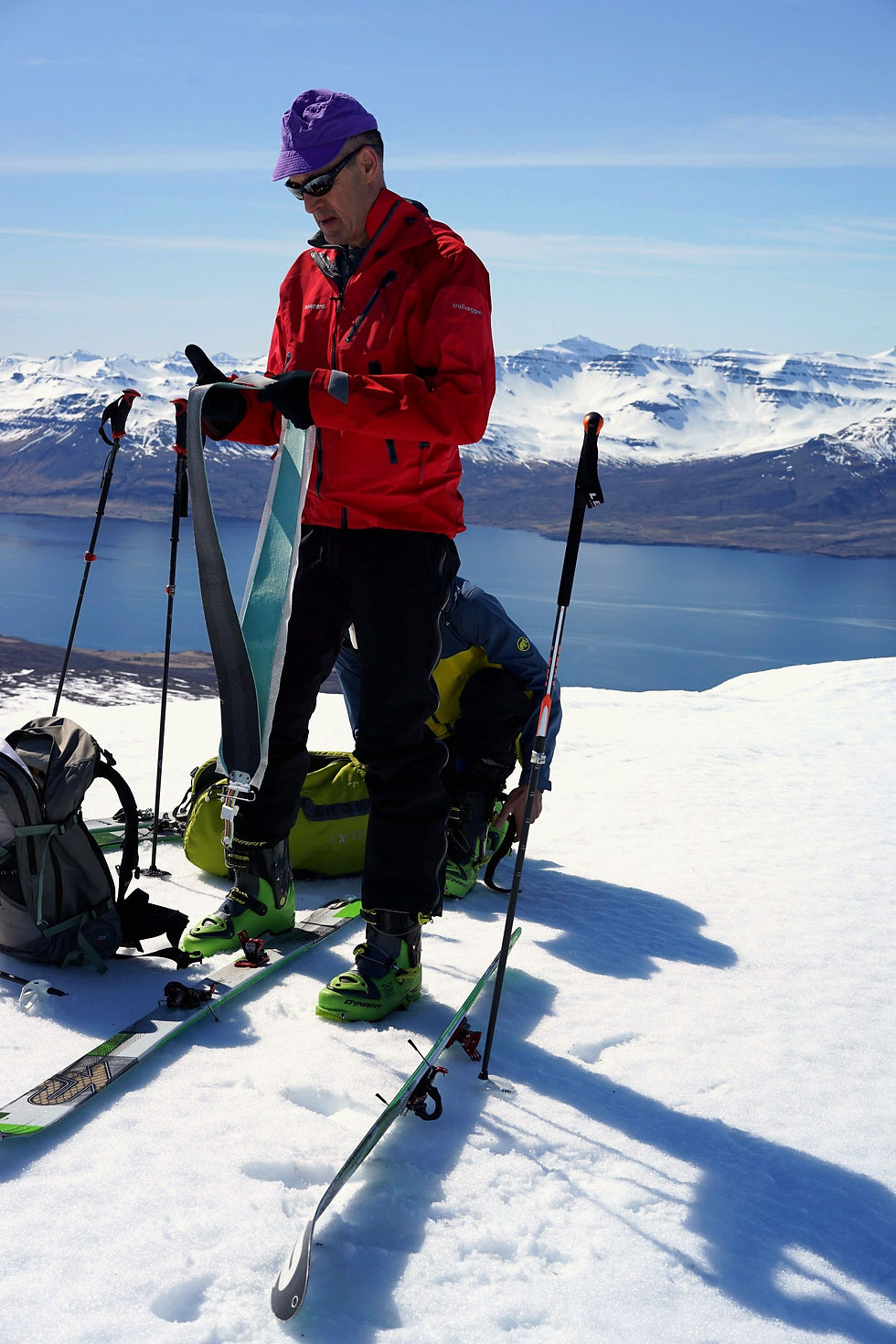 Un skieur range sa peau de phoque au sommet, sacs à dos et skis posés dans la neige, un fjord derrière lui