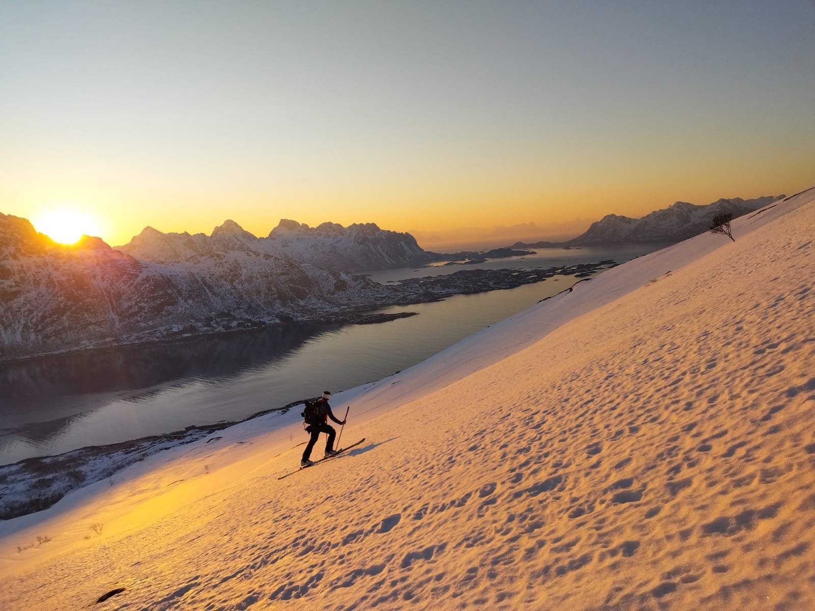 Ein einzelner Skifahrer quert ein weites, von der tiefen Sonne golden gefärbtes Schneefeld, darunter der Fjord und die Lofoten