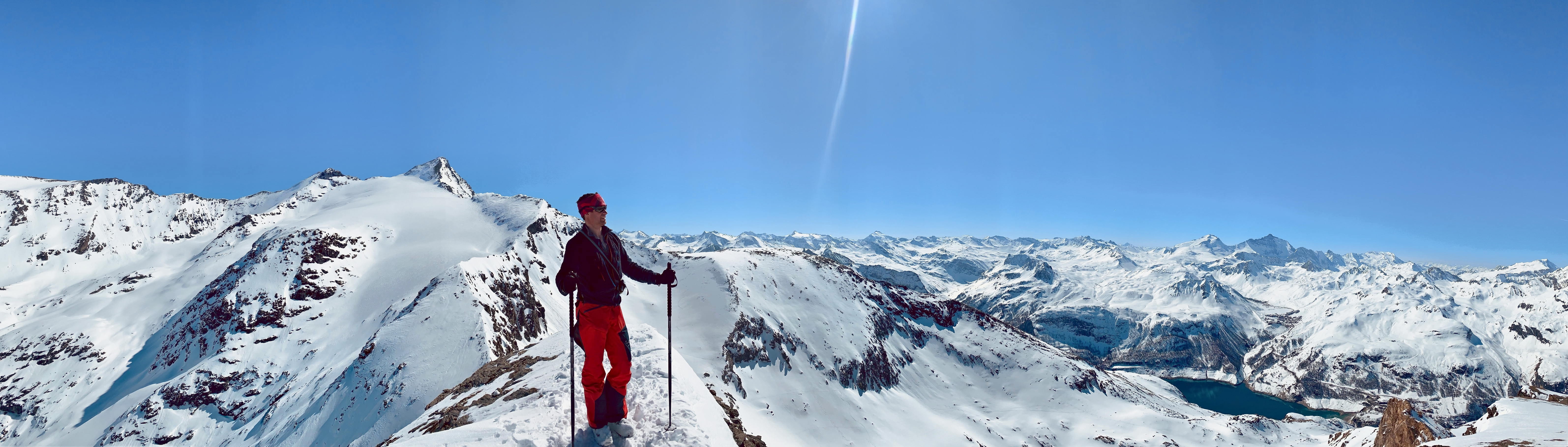 A skier standing on a ridge above a panorama of snowy peaks and the Lac du Chevril reservoir