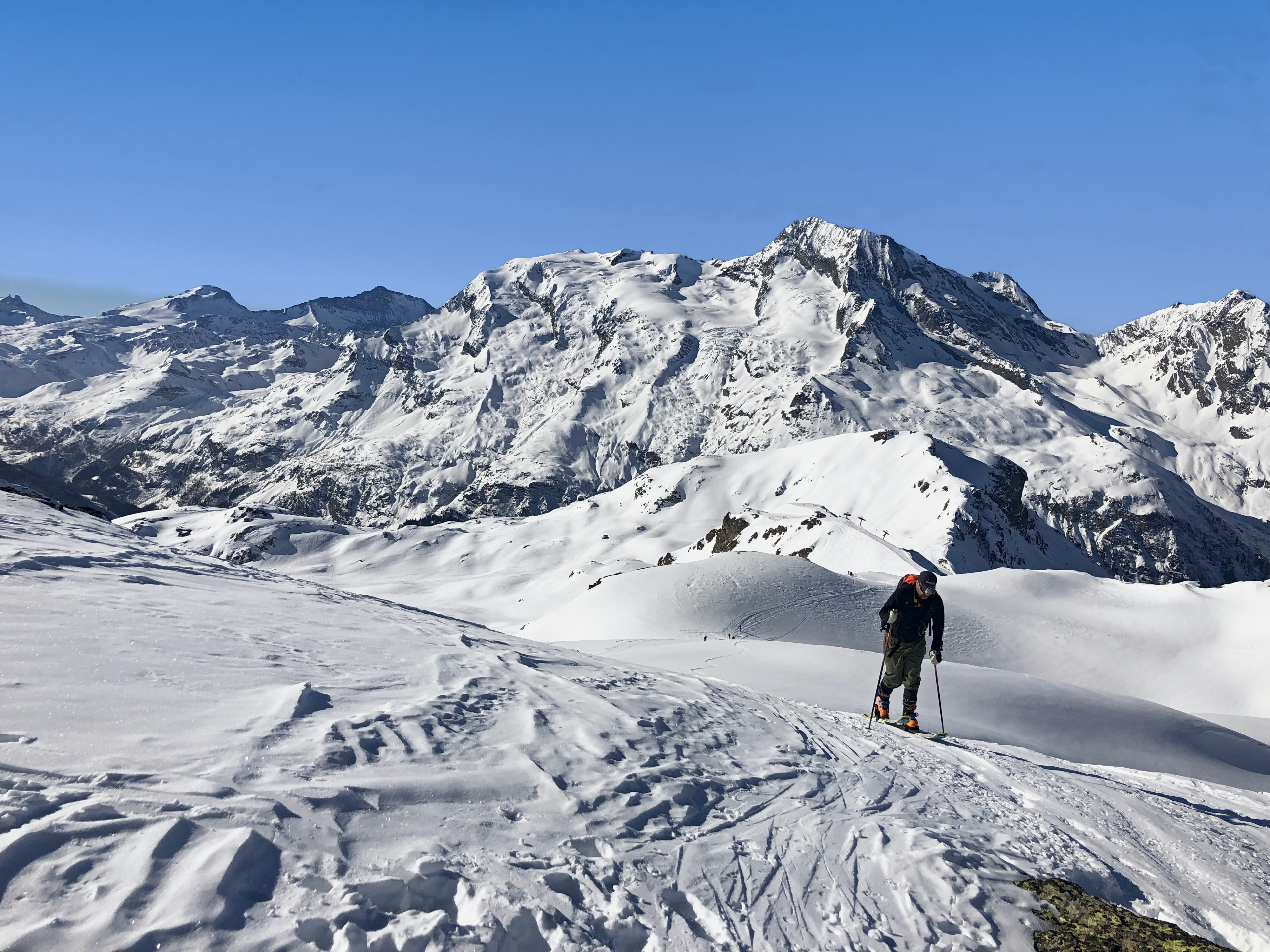 Lone ski tourer skinning across a snowy shoulder facing a big glaciated massif