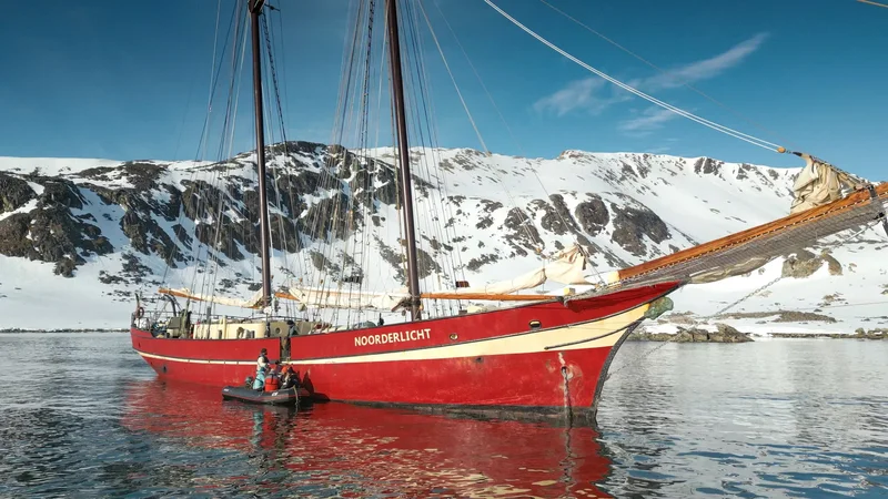 The red schooner Noorderlicht at anchor below snow-covered Spitsbergen mountains, her tender alongside