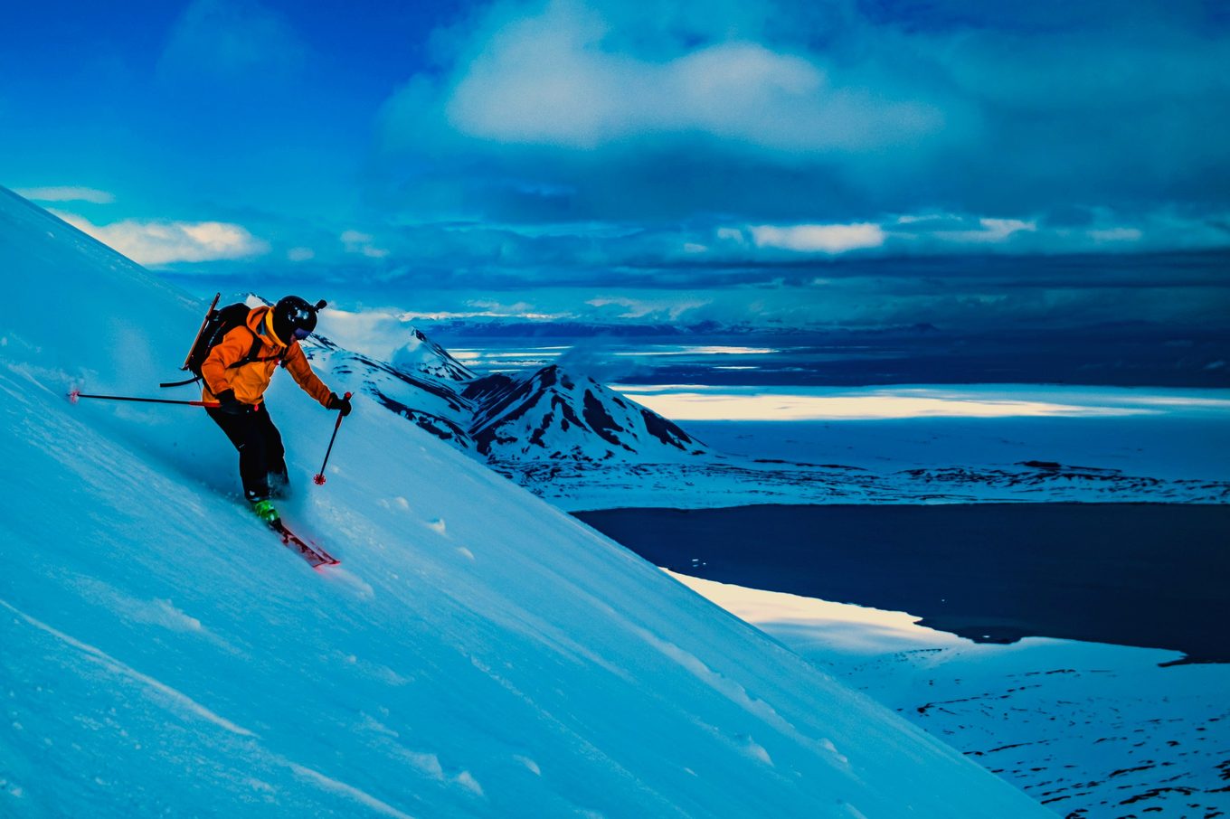 A skier carves a steep slope above an Arctic fjord in blue end-of-day light.