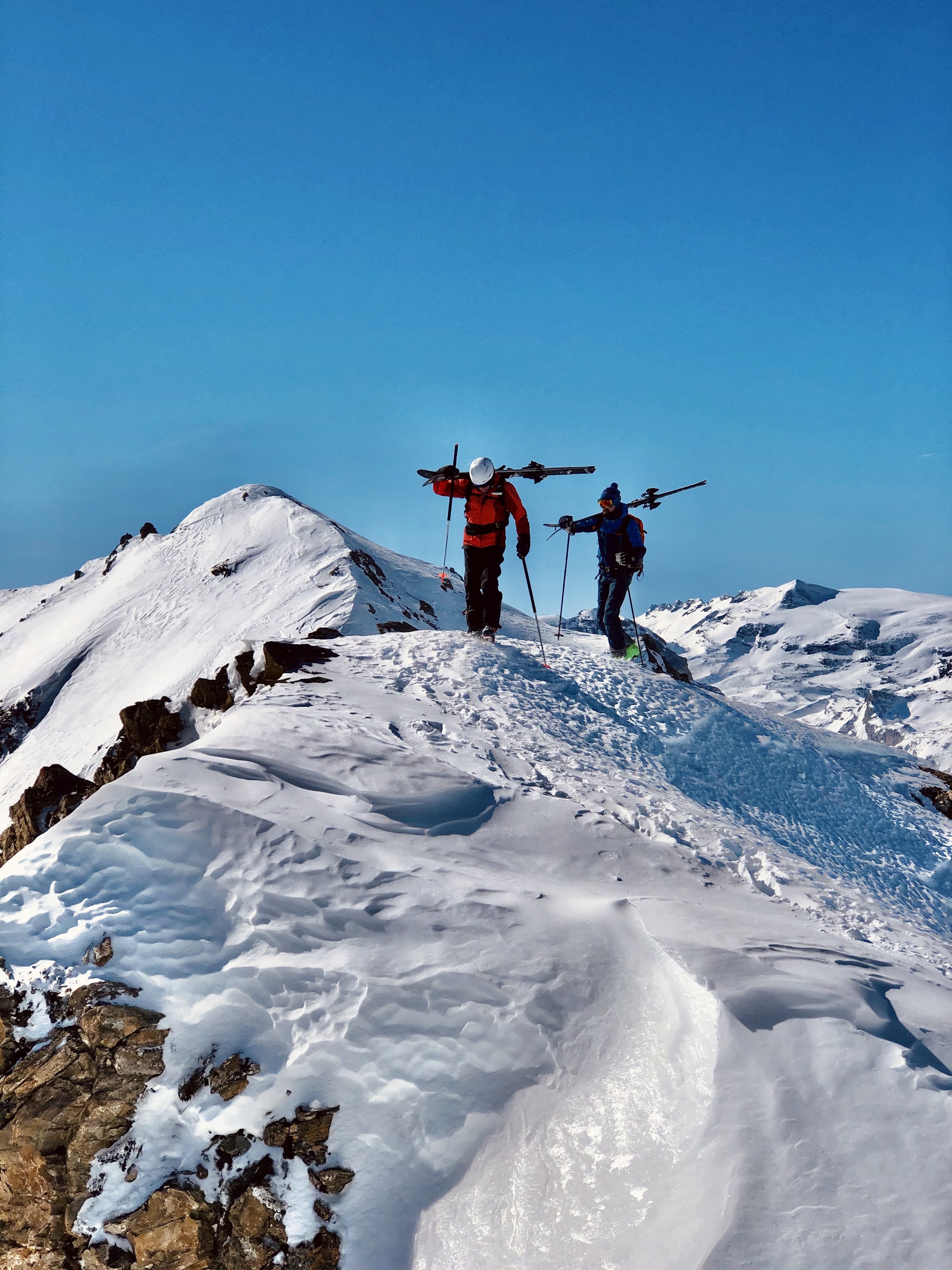Zwei Skifahrer auf einem verschneiten Grat, die Ski geschultert, unter blauem Himmel
