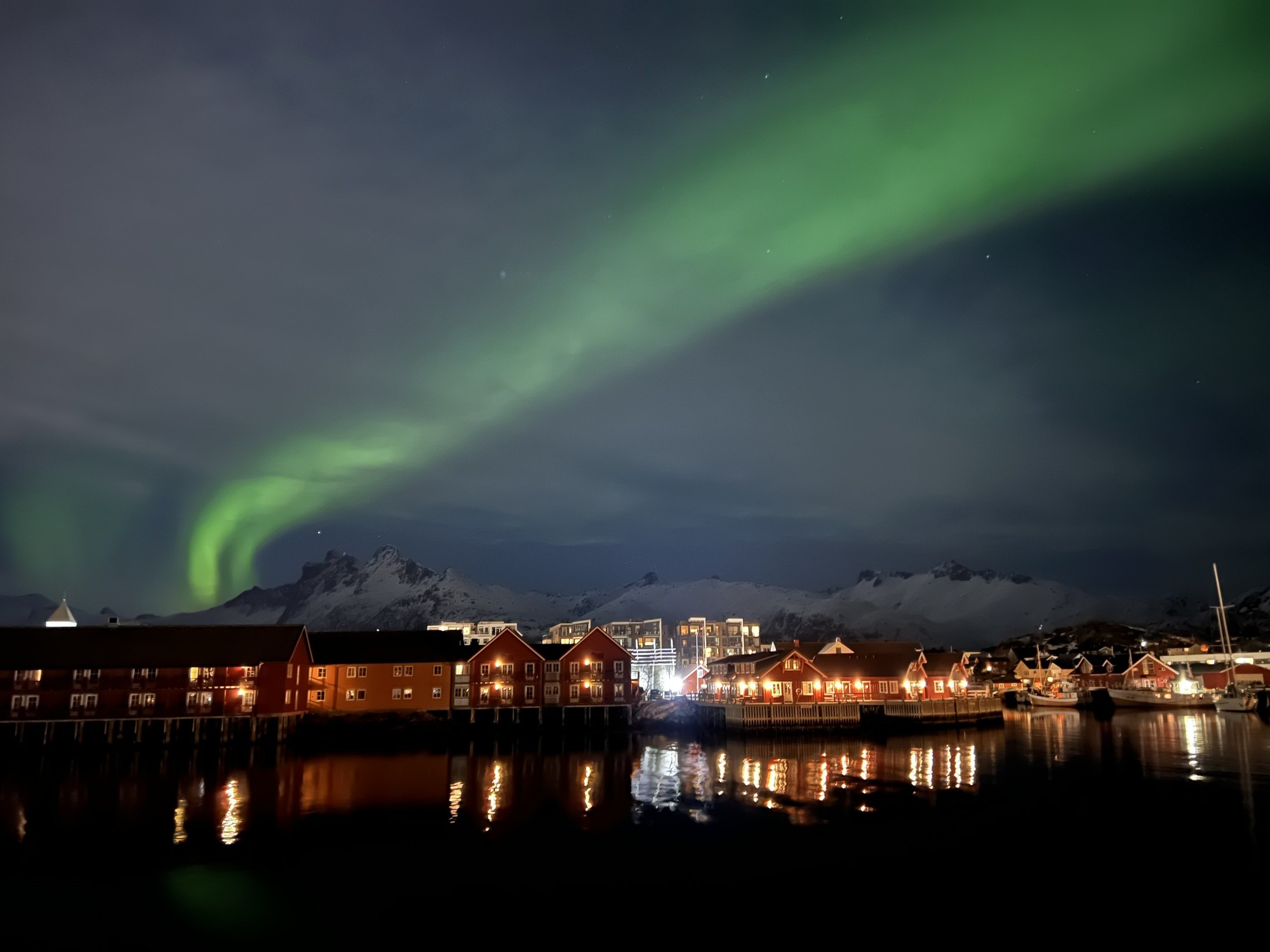 Ein grünes Nordlicht über der Uferfront von Svolvær, Holzhäuser mit erleuchteten Fenstern und Spiegelungen auf dem schwarzen Wasser
