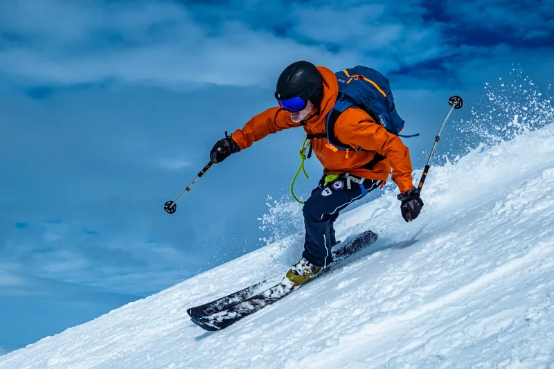 A skier in an orange jacket in Spitsbergen powder, a plume of snow against a blue sky