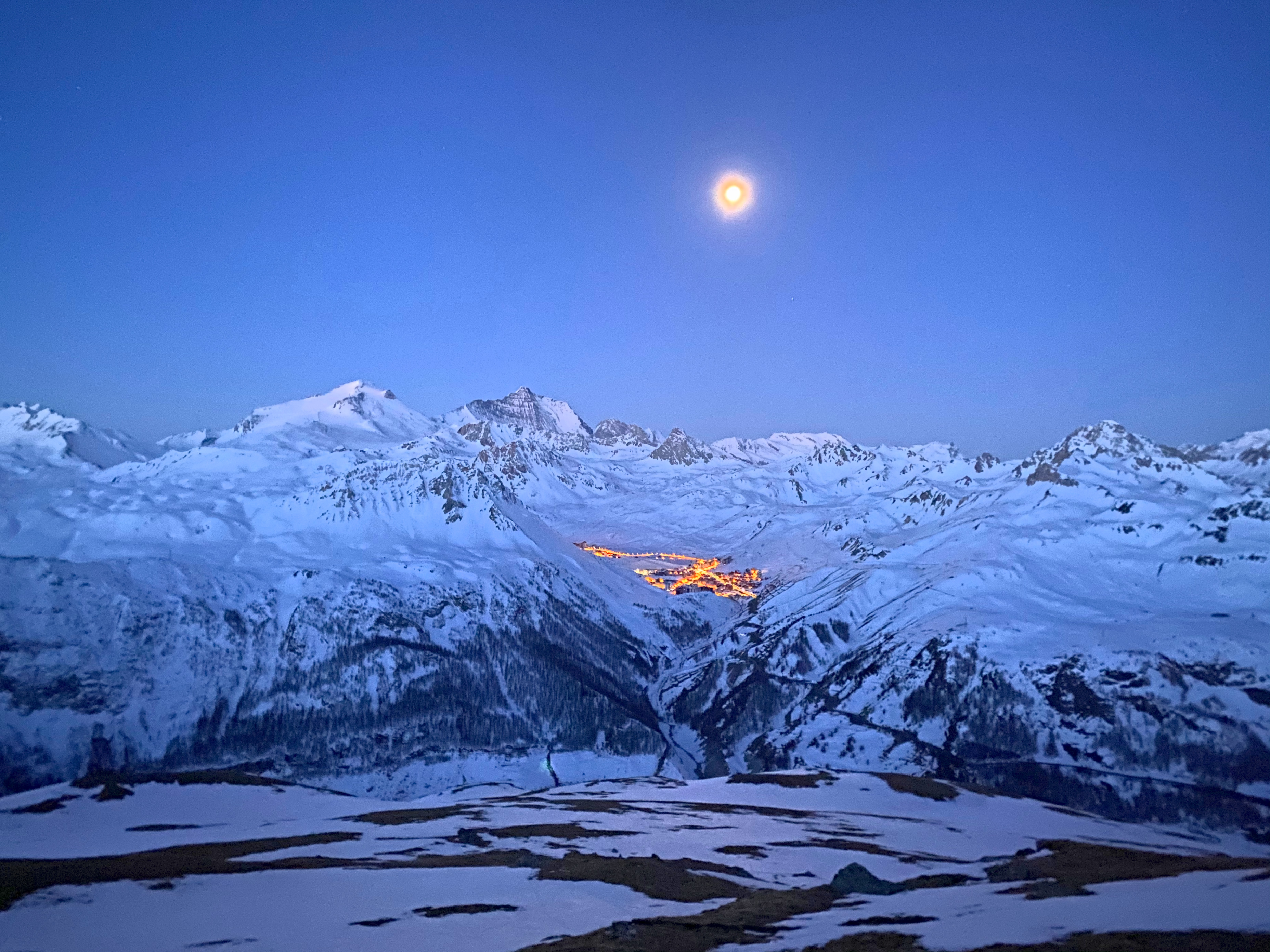 Schneesicherheit in Val d'Isère: das beleuchtete Dorf im verschneiten Tal zur blauen Stunde, der Mond über den Gipfeln
