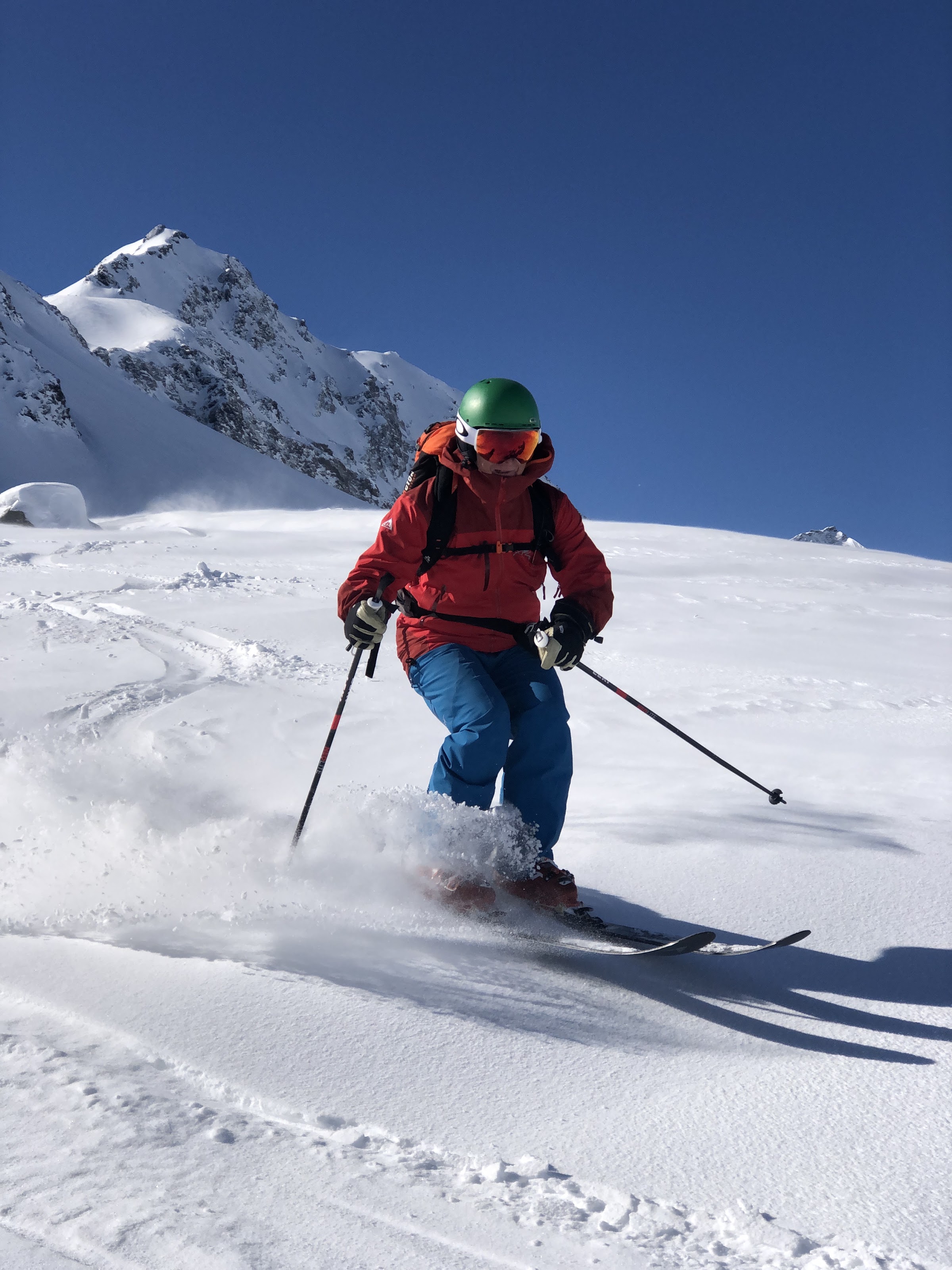 Skifahrer in roter Jacke und grünem Helm im Pulverschnee unter einem Felsgipfel und blauem Himmel