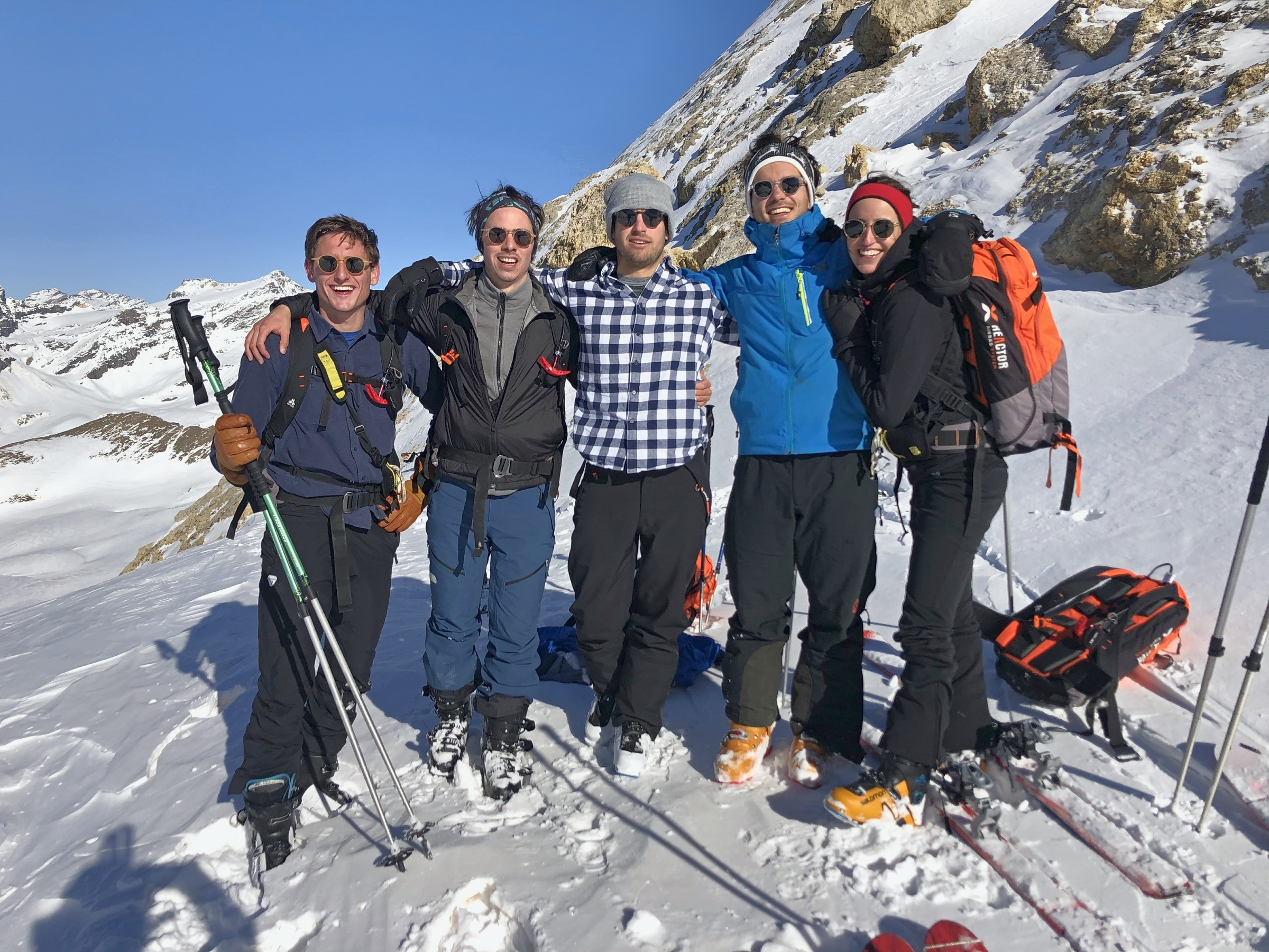 Five smiling ski tourers arm in arm in the sun, high in the snowy mountains