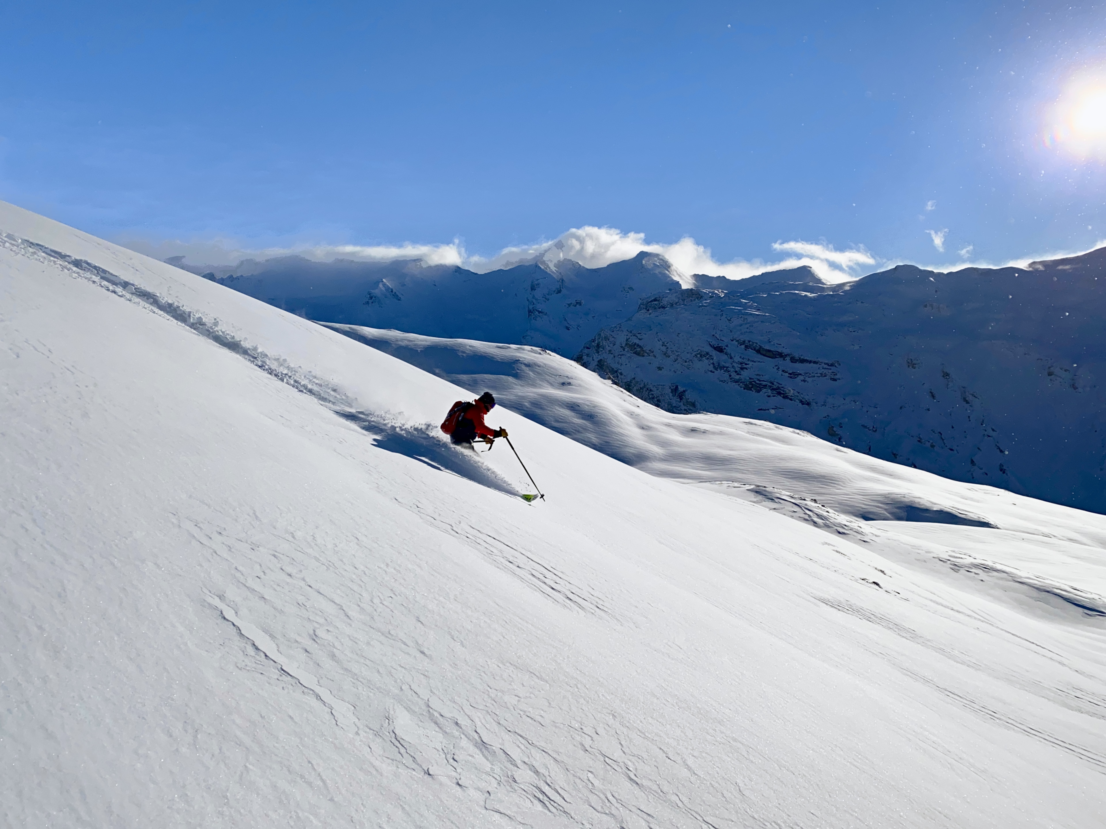 Heliskiing: Skifahrer in roter Jacke im großen Pulverhang, dahinter ein verschneites Massiv und tiefe Sonne
