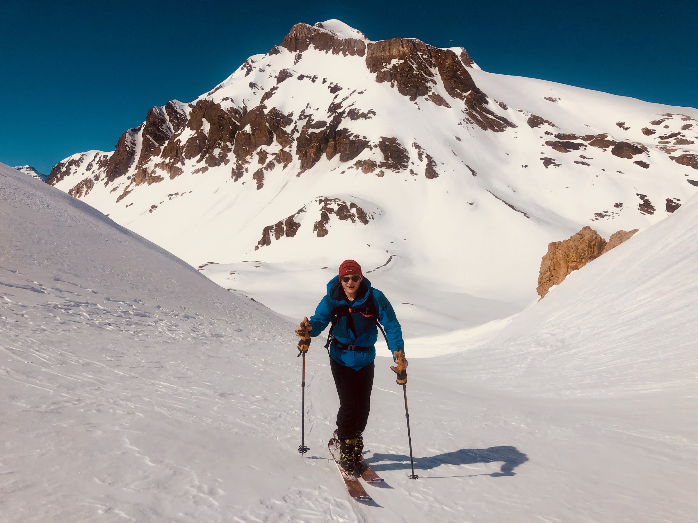 Skier in a blue jacket skinning up a snowy valley below a rocky summit