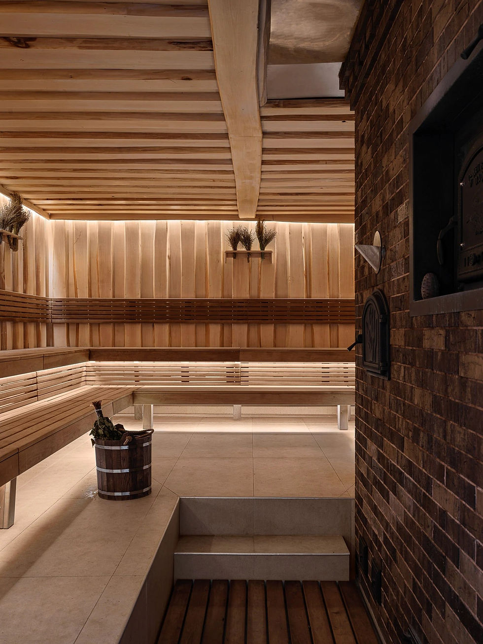 Intérieur d'une banya : gradins de tilleul, mur de briques, porte de poêle en fonte et seau de chêne avec un bouquet de bouleau