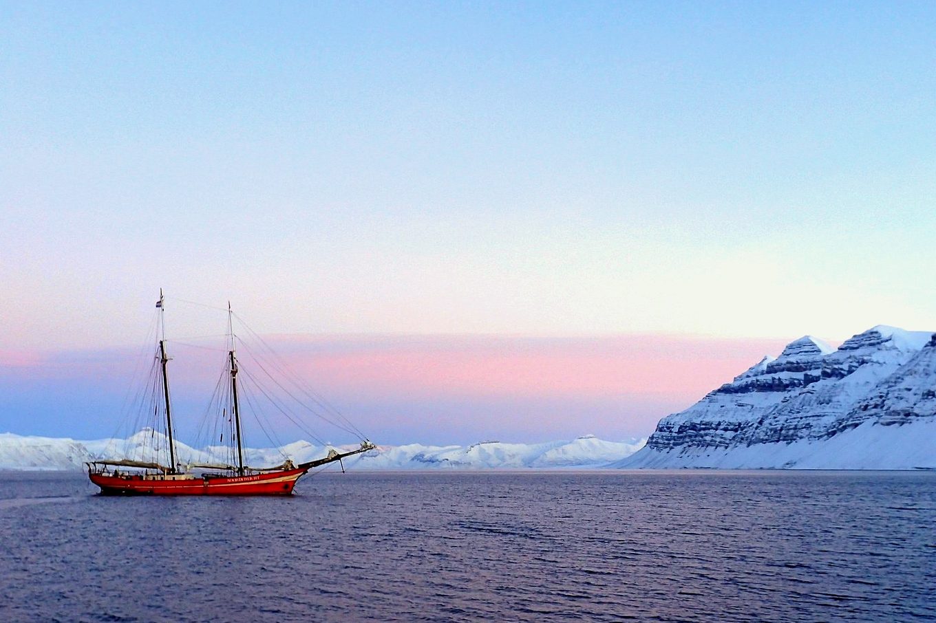 A red schooner at anchor in a calm fjord under a pink polar-night sky, snowy mountains on the horizon.