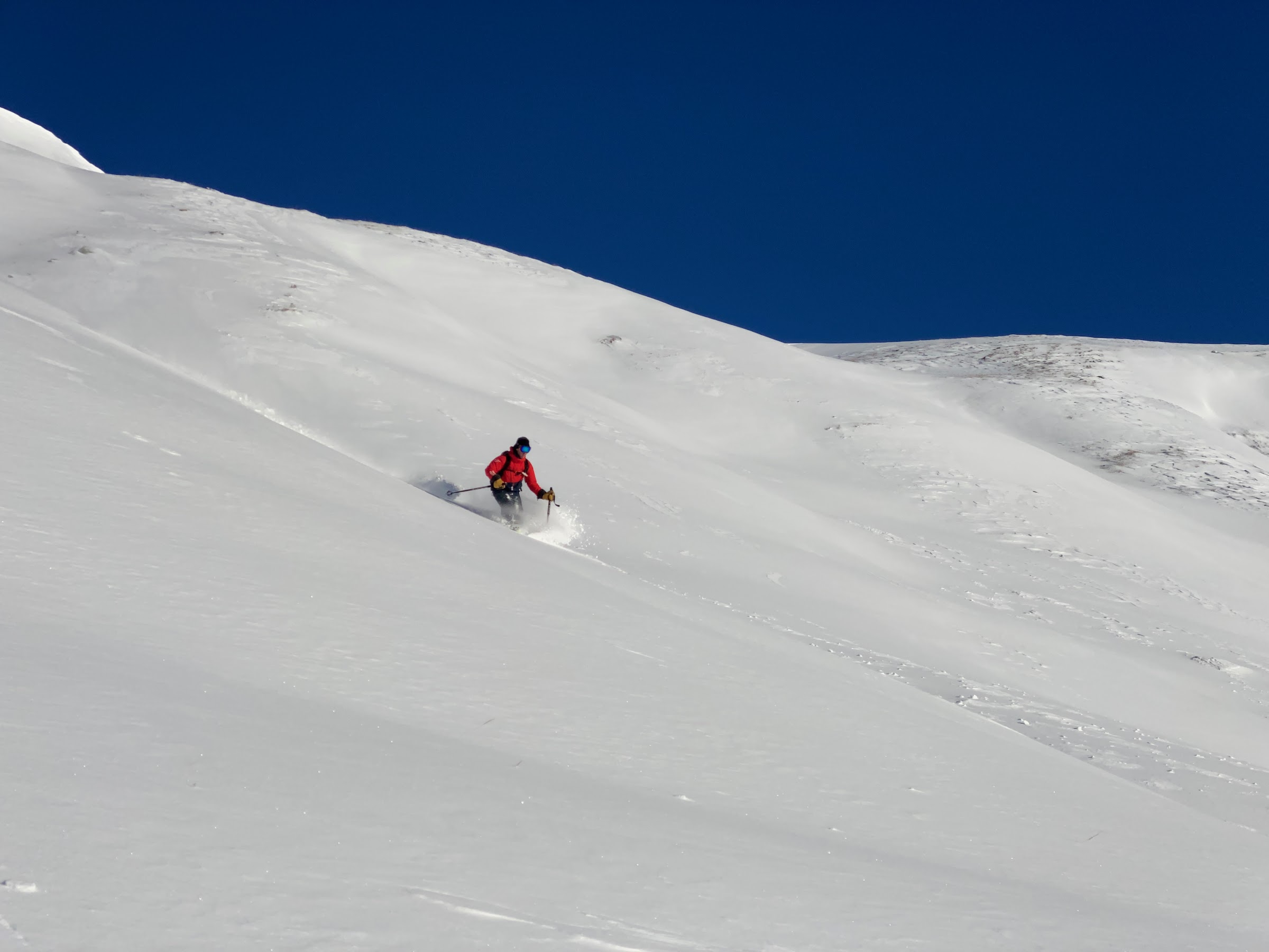 Einzelner Skifahrer in roter Jacke in einem riesigen unverspurten Hang unter tiefblauem Himmel