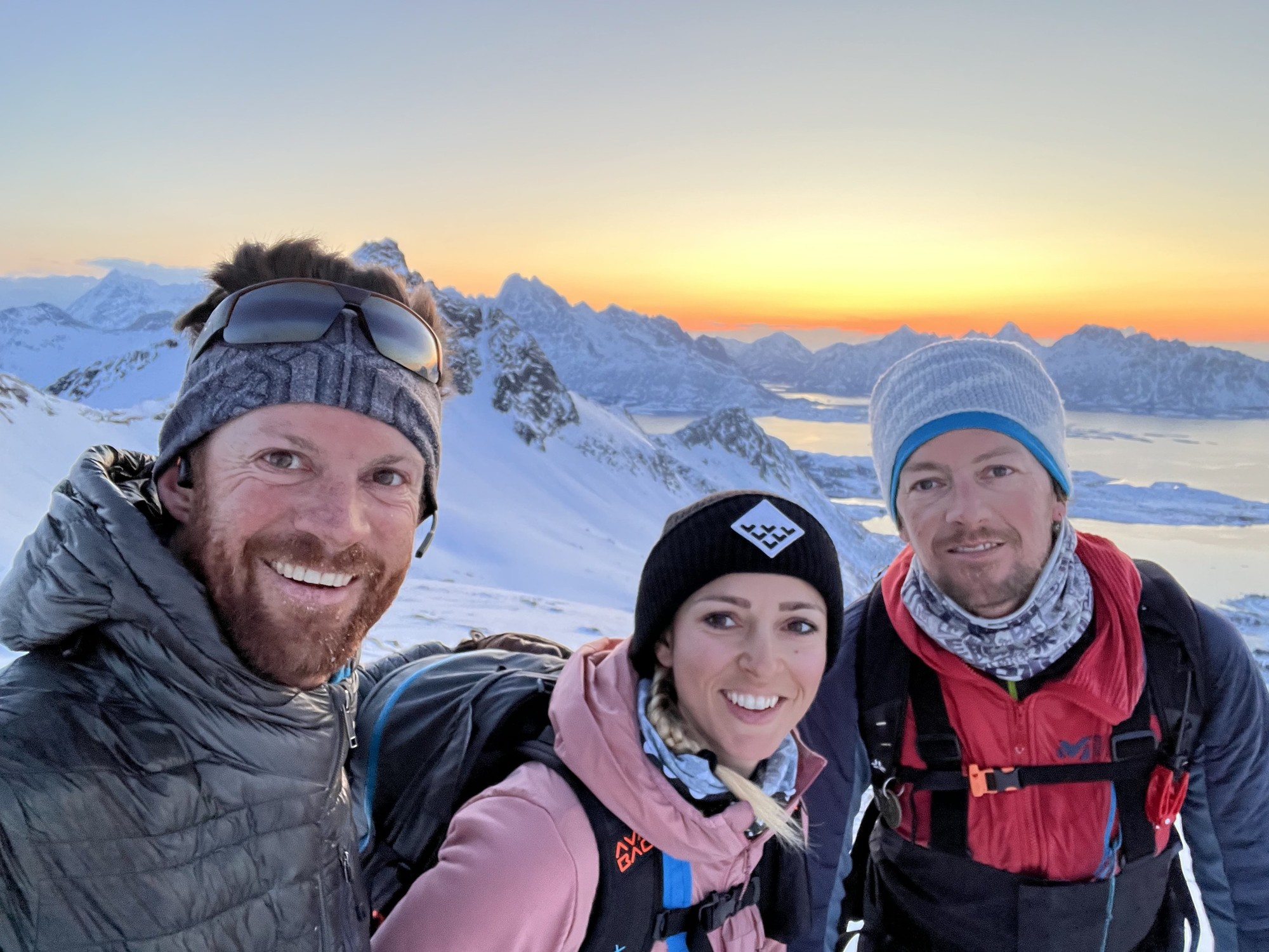 Drei Skifahrer zusammen am Gipfel, lachend, der orange Abendhimmel und das Meer zwischen den Inseln