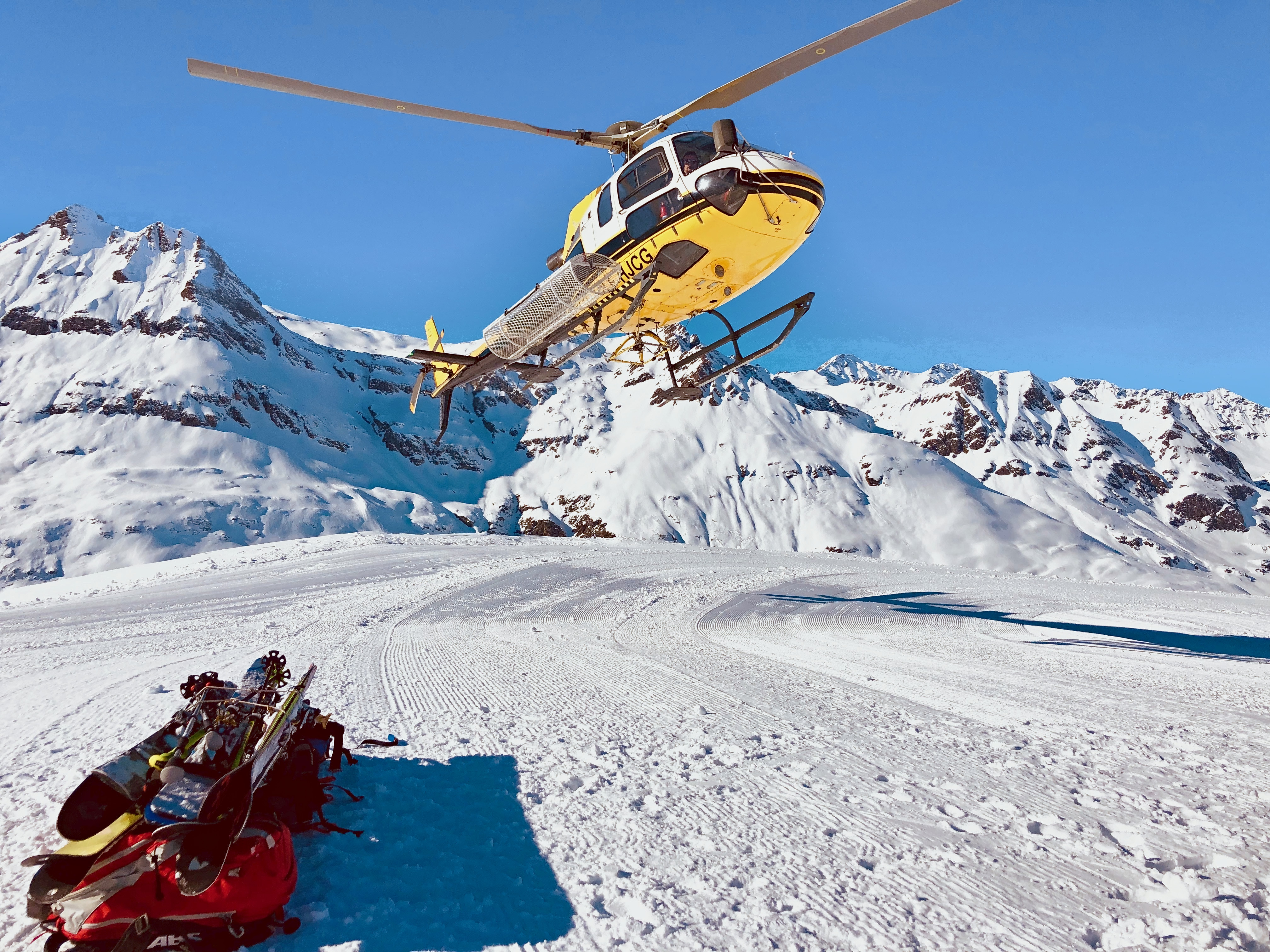 Heliskiing: gelber Hubschrauber über einem verschneiten Landeplatz vor den Gipfeln, Ski und Lawinenairbag-Rucksack im Schnee