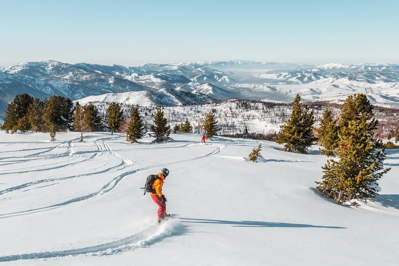 Un snowboardeur en veste orange ride de larges courbes, panorama de l’Altaï sous un ciel bleu clair
