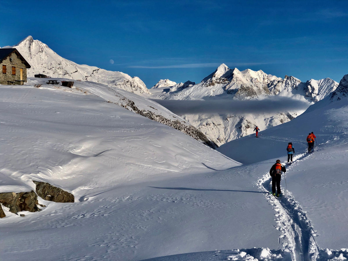 Small group skinning up a track towards a stone hut, snowy peaks behind