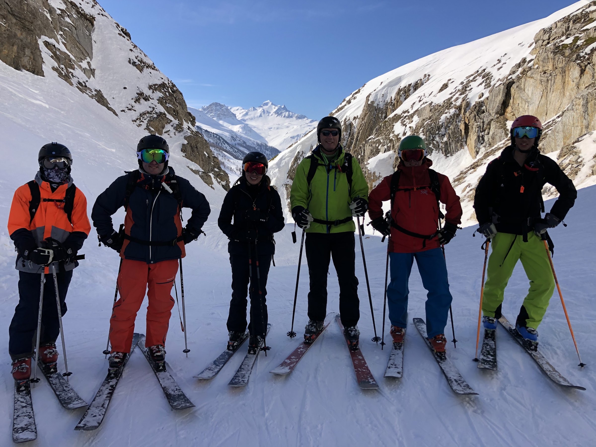 Six skiers in helmets standing side by side in a narrow valley, snowy peaks behind