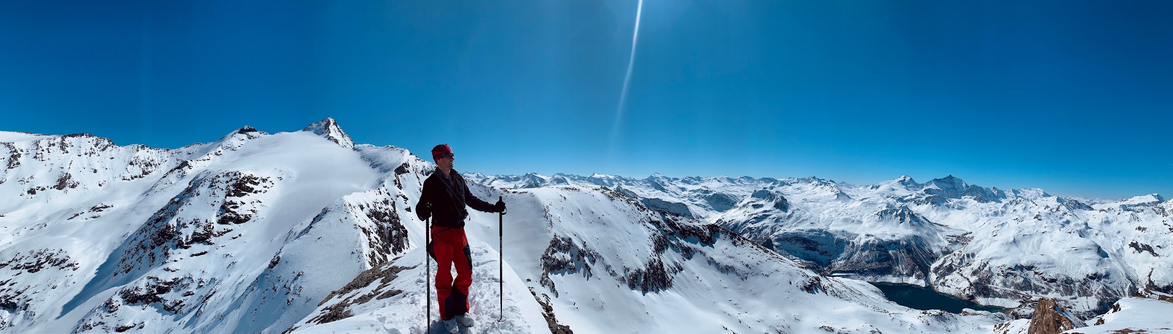 Skifahrer auf einem Grat über einem Panorama verschneiter Gipfel und dem Stausee Lac du Chevril