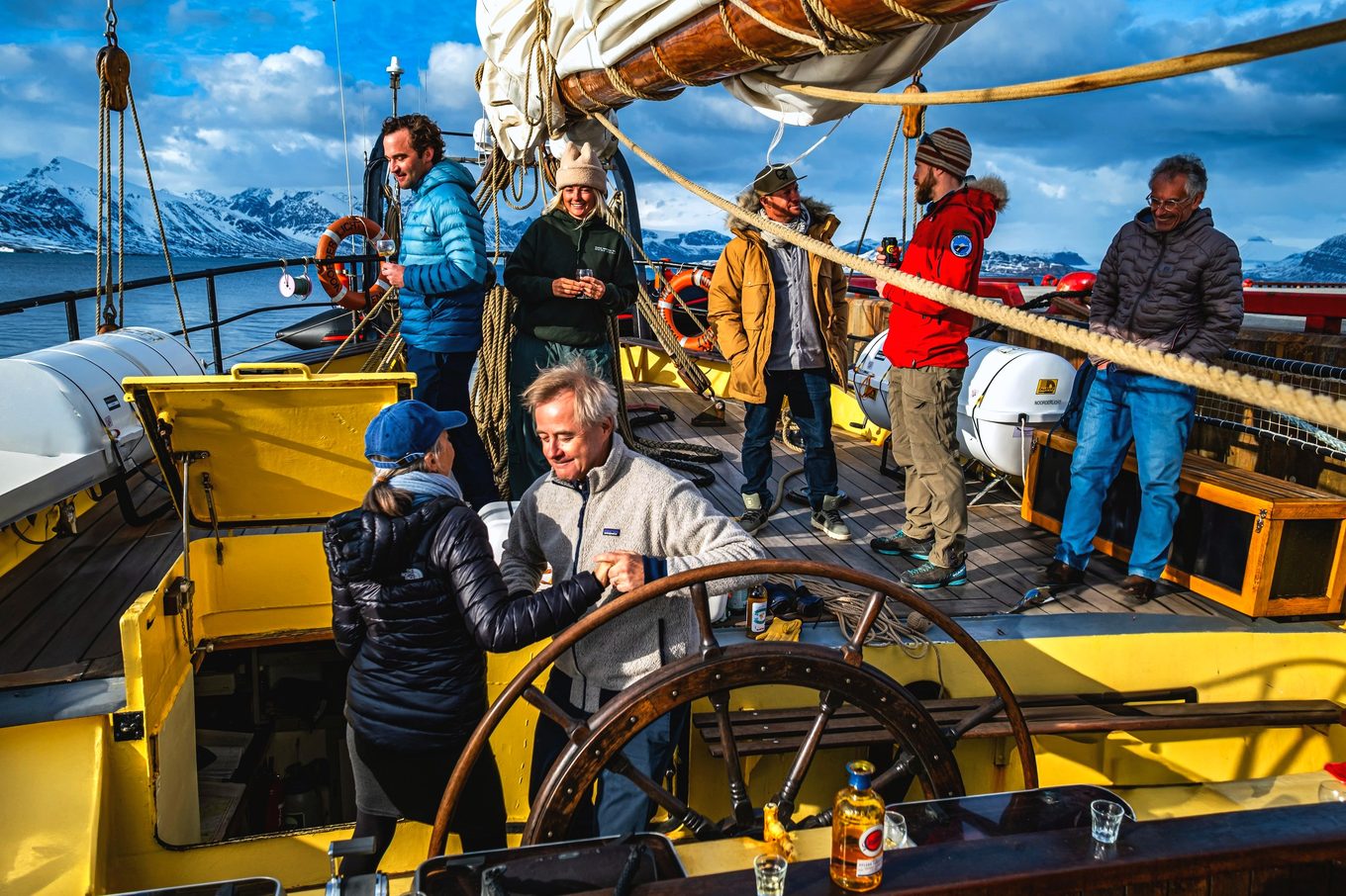A group gathers on the wooden deck of a schooner in low golden light, snowy mountains behind.