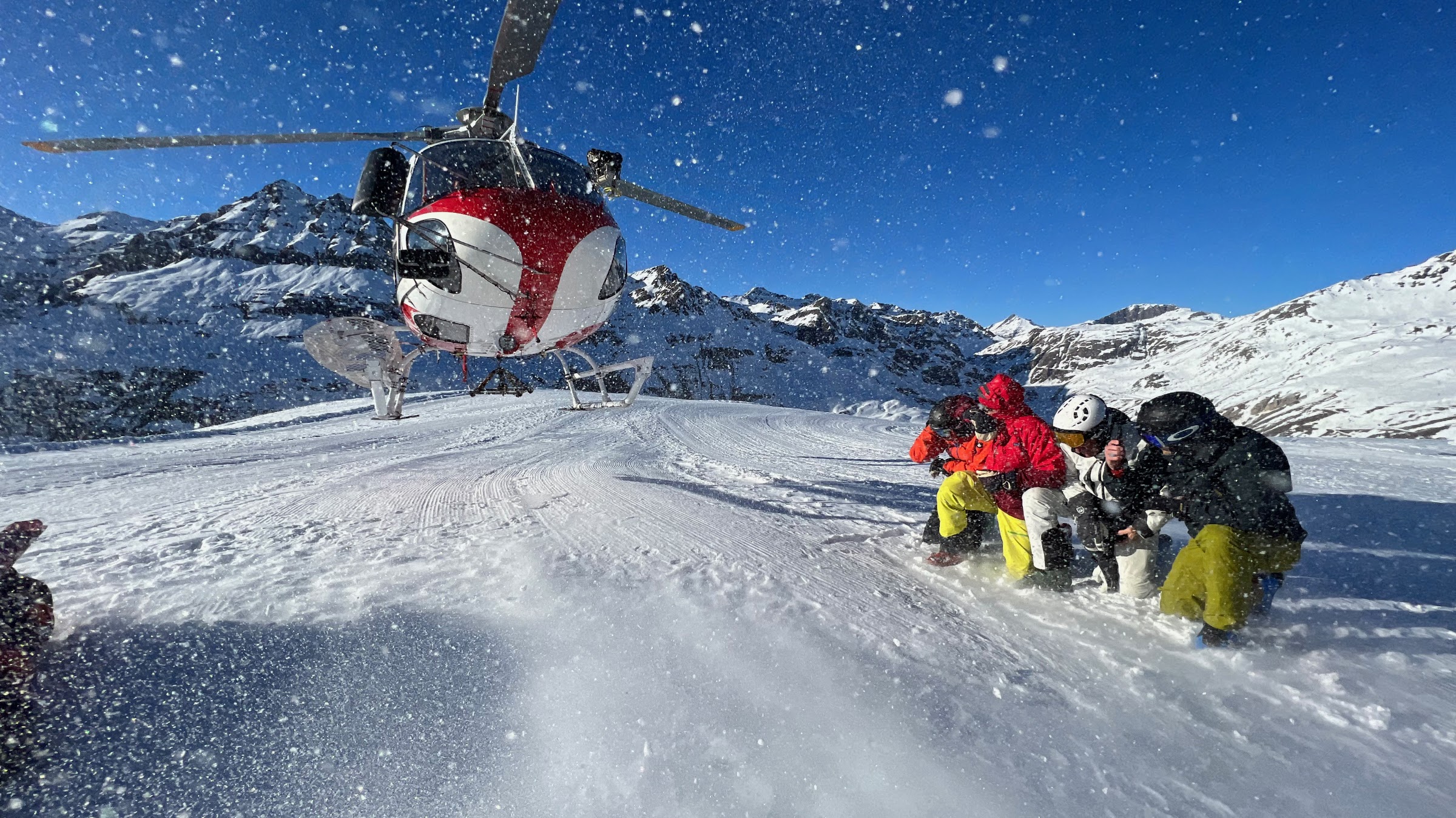 Hubschrauber mit laufendem Rotor im aufgewirbelten Schnee, Skifahrer hocken am Rand des Landeplatzes