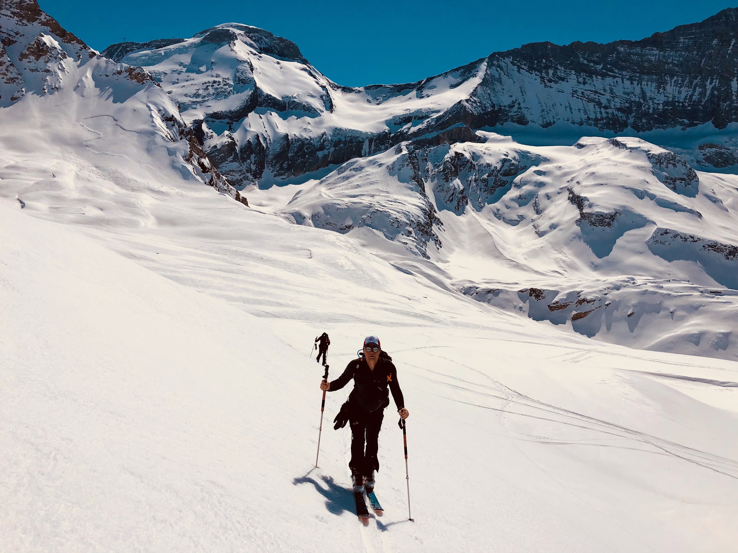 Skitourengeher beim Aufstieg mit Fellen am weiten Hang, ein zweiter weiter oben, dahinter ein verschneites Massiv