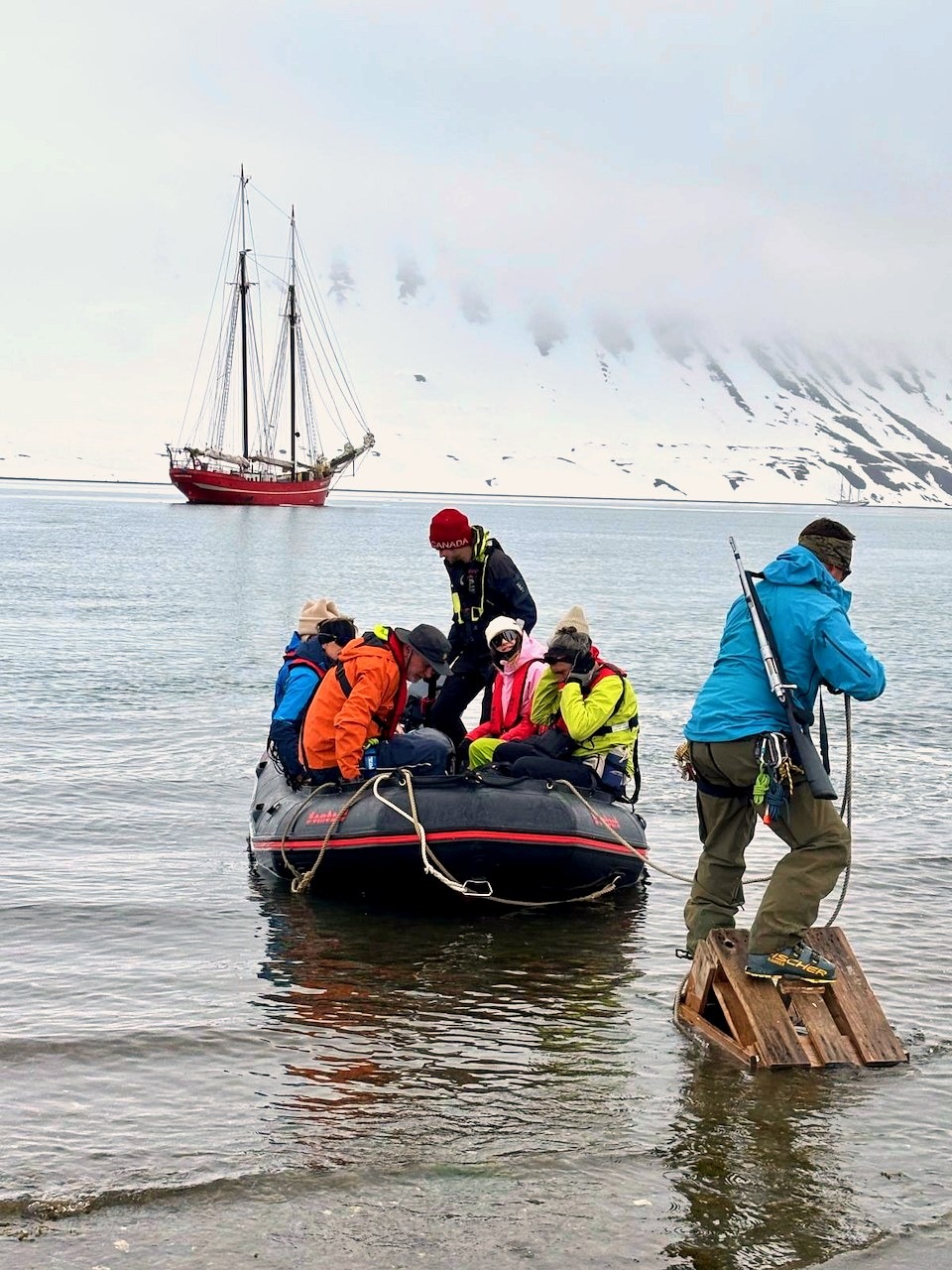 Skiers step off a zodiac at the foot of a snowy mountain, a schooner at anchor behind in the mist.