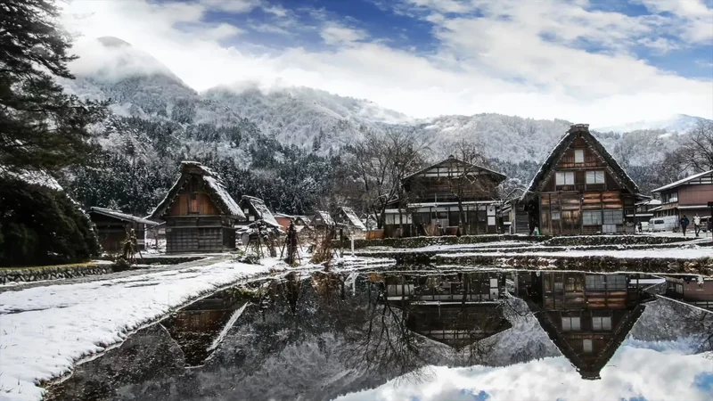 In Japan skifahren: traditionelles Dorf mit Strohdächern im Schnee, das sich in einem Wasserbecken spiegelt, dahinter verschneite Berge