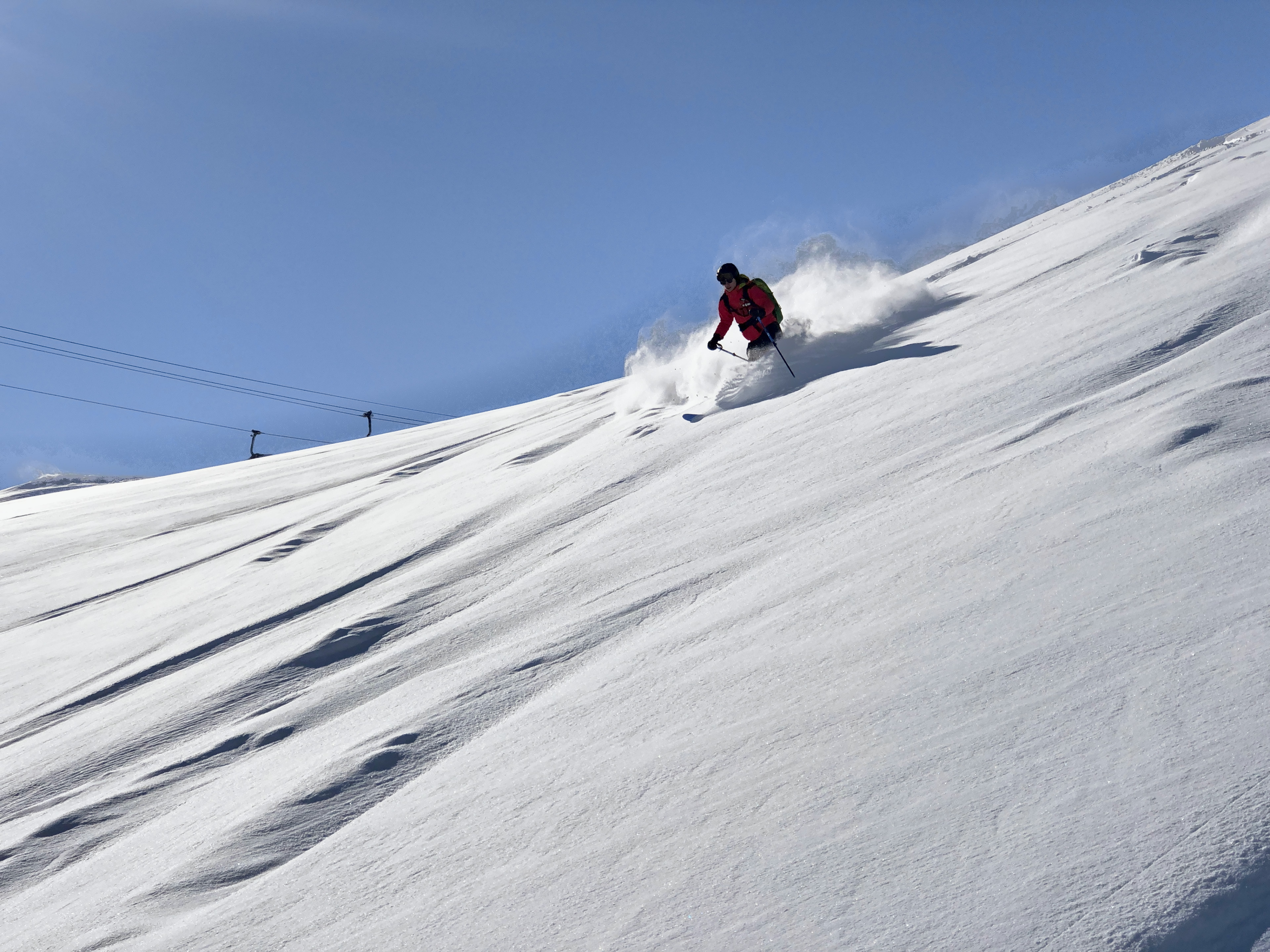 Tiefschneefahren: Skifahrer in roter Jacke wirbelt am Steilhang Pulverschnee auf, in der Ferne ein Skilift