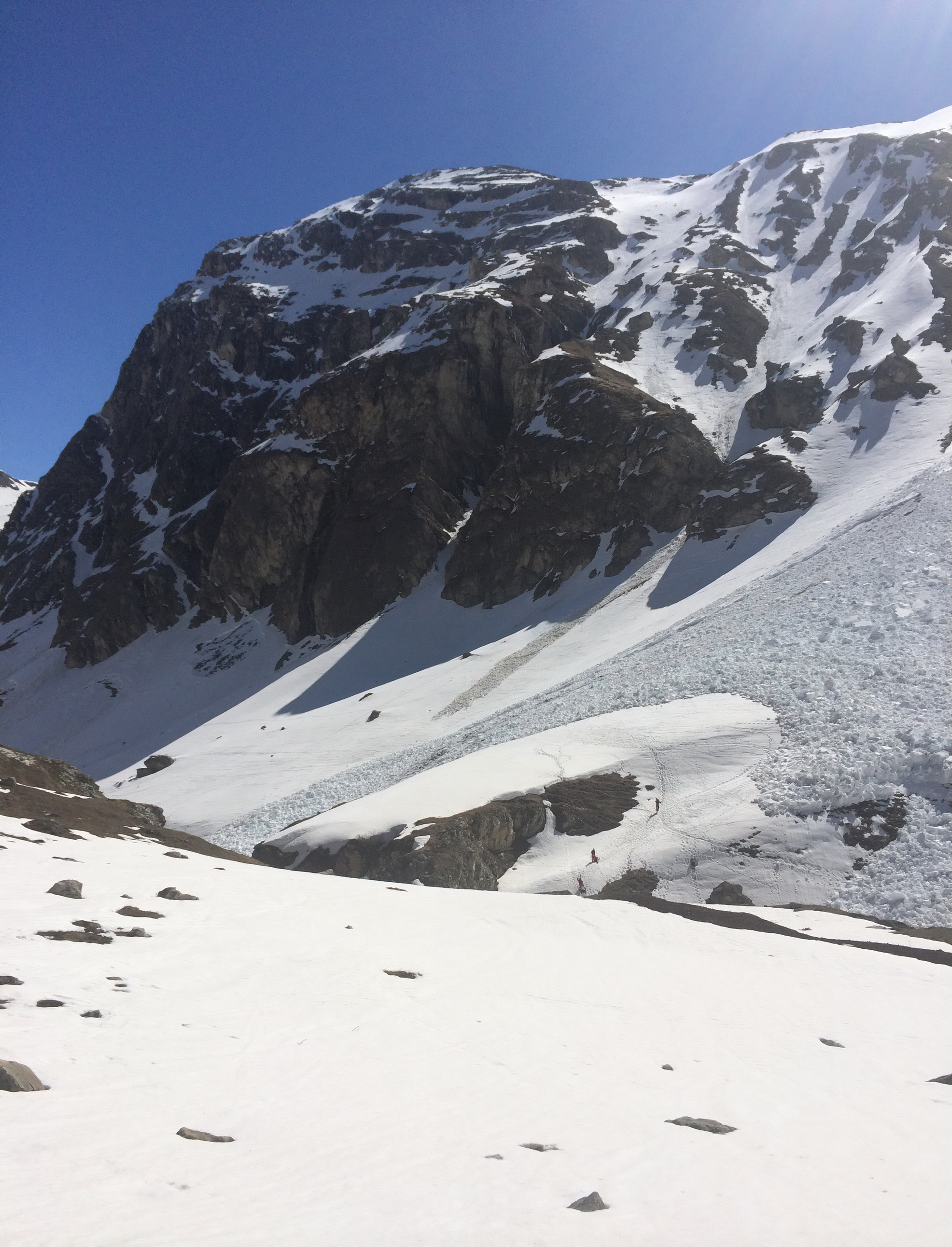 Lawinenablagerung aus großen Schneeblöcken am Fuß einer Felswand bei strahlendem Wetter, daneben einige winzige Gestalten
