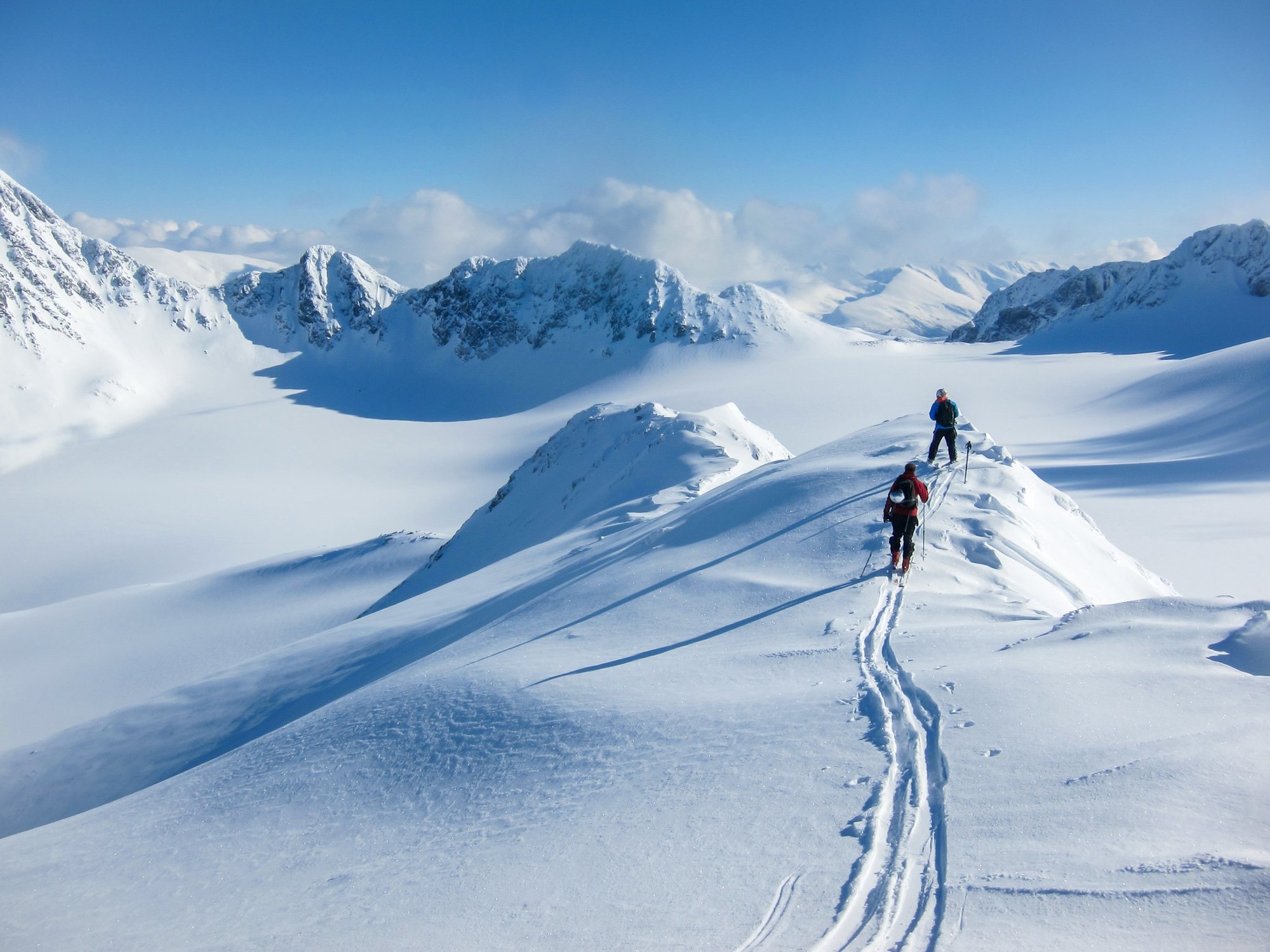 Eine lange Zickzack-Aufstiegsspur auf Fellen über einen unverspurten, vom Wind bearbeiteten Hang, zwei Skifahrer erreichen den Grat