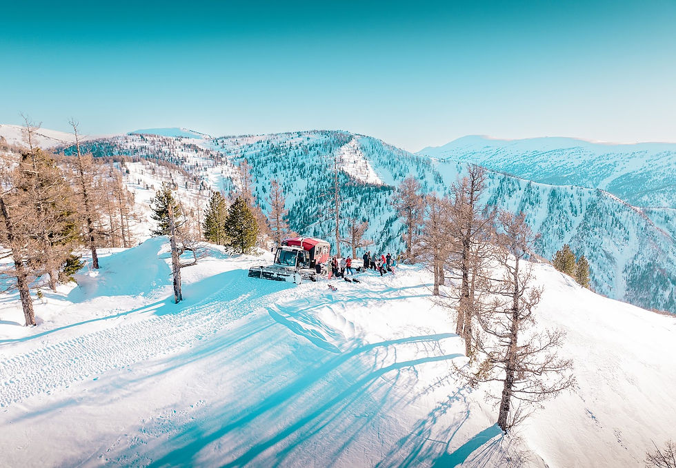 Groupe de skieurs rassemblés autour d'une chenillette rouge sur une croupe, vallées boisées de l'Altaï en arrière-plan