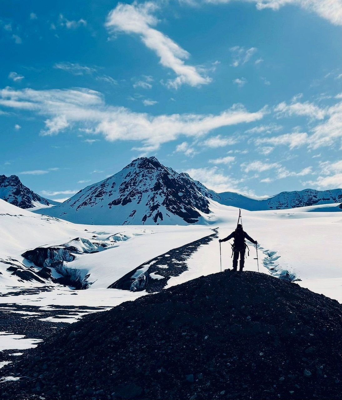 A skier, skis on pack, looks out at a sharp peak above an Arctic glacier.