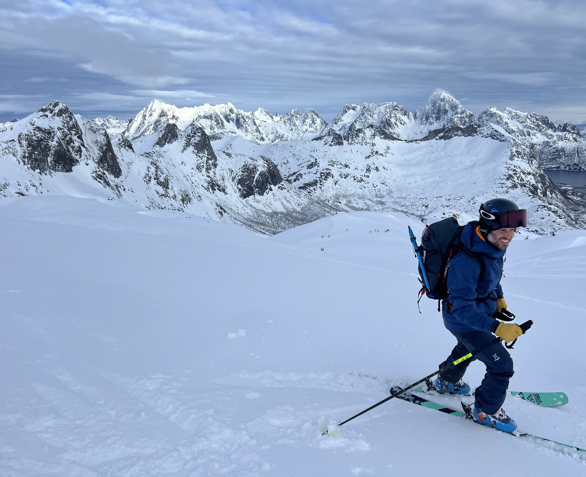 Ein lächelnder Skifahrer über einem weiten Schneefeld, die zerklüfteten Lofoten-Grate unter tiefer Wolkendecke, rechts der dunkle Fjord