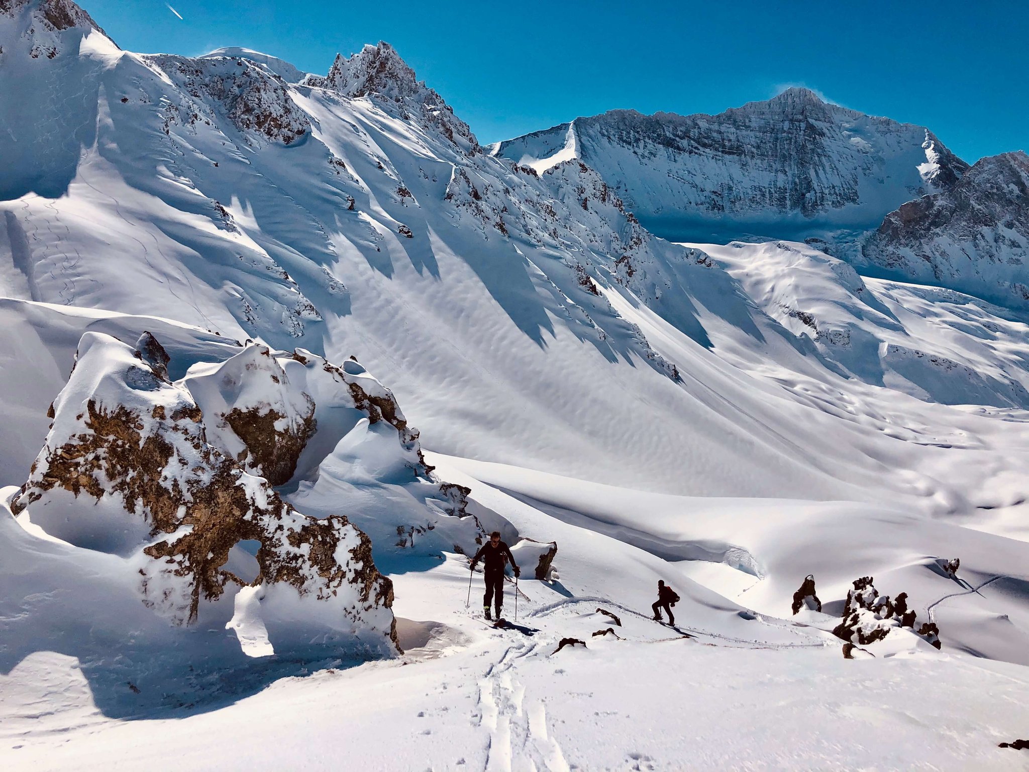 Zwei Skitourengeher beim Aufstieg mit Fellen zwischen verschneiten Felsen bei Val d'Isère, große Wand dahinter