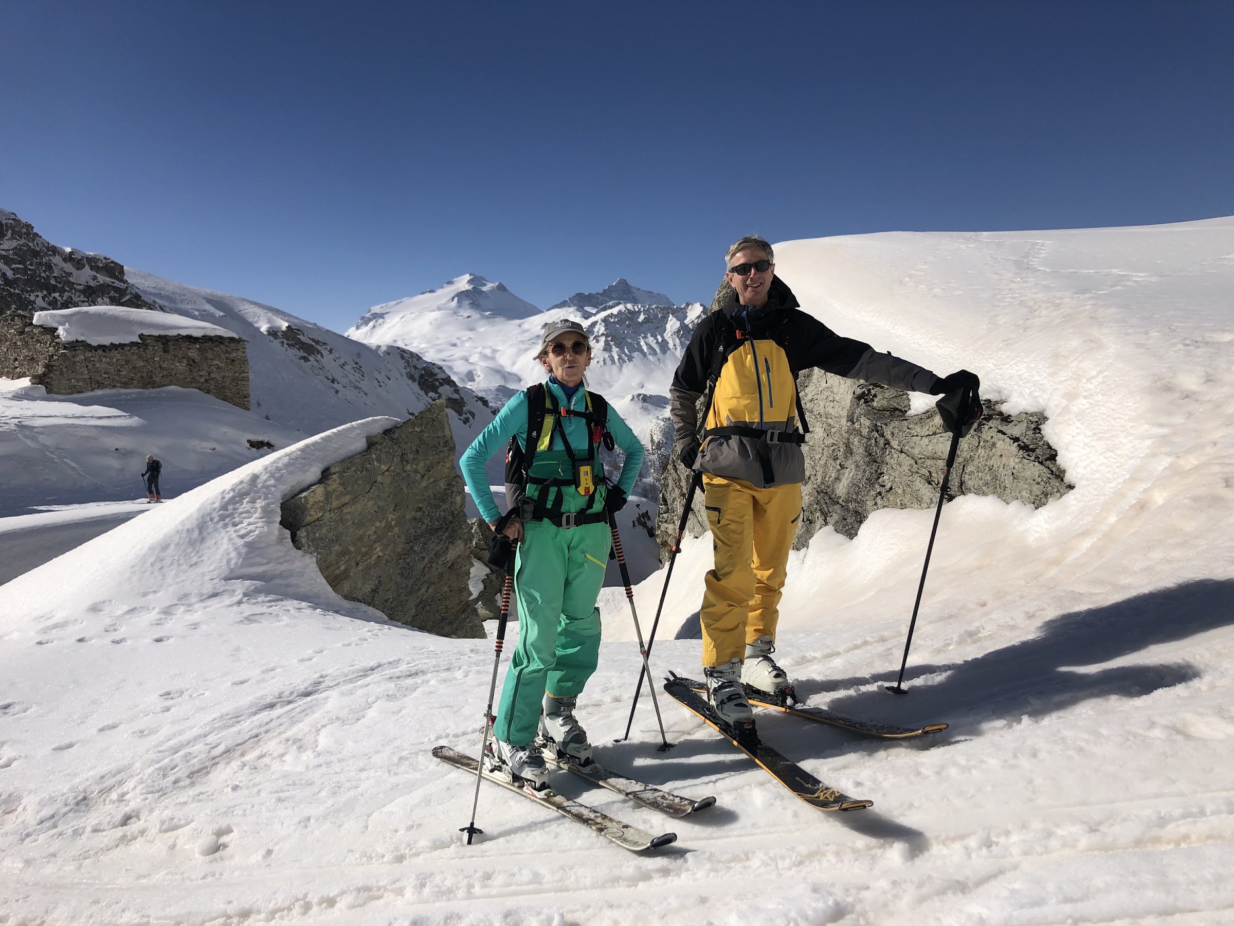 Two smiling skiers with backpacks pose in the sun beside a snow-capped boulder