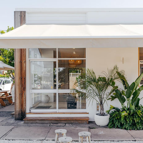 Modern white building with large glass windows, green plants, and outdoor stools.