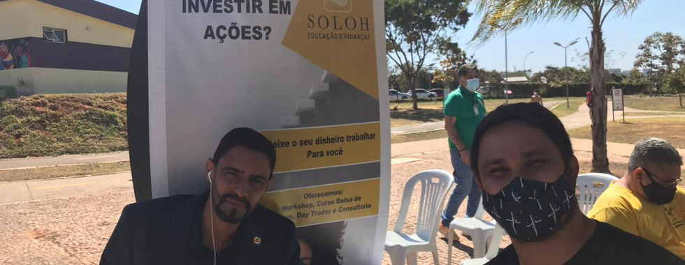 Two people at an outdoor booth, "Já pensou investir em ações?" banner