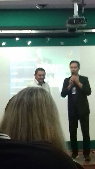 Two men presenting; one in a suit, another in a white shirt; audience visible
