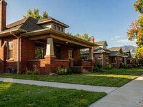 Sunlit brick houses on a quiet tree-lined suburban street with sidewalks, green lawns, and mountains in the distance.