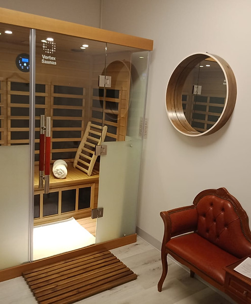 Wooden infrared sauna unit with glass front. Set up with wooden backrest, white towel rolled up on bench and white footmat. Circular wooden mirror on wall. Red vintage ornate bench chair below mirror