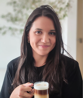 Imagem de Bia Bonas, sorrindo, cabelos pretos, morena, com um copo de café na mão, cabelos escuros divididos ao meio.