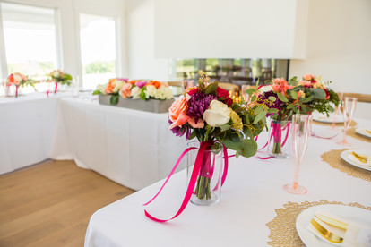 Inside the Selah Event Venue ballroom three long tables are decorated with white tablecloths, gold place settings, white plates, and golden utensils. At the head table placed before the room's fireplace, is a long bouquet of colorful flowers. Image captured by Photography of Sarah Mae.
