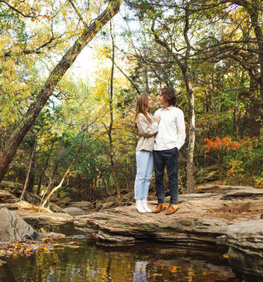 A man and woman stand on the edge of a large stone hanging over a creek. In the water below them is their reflection.