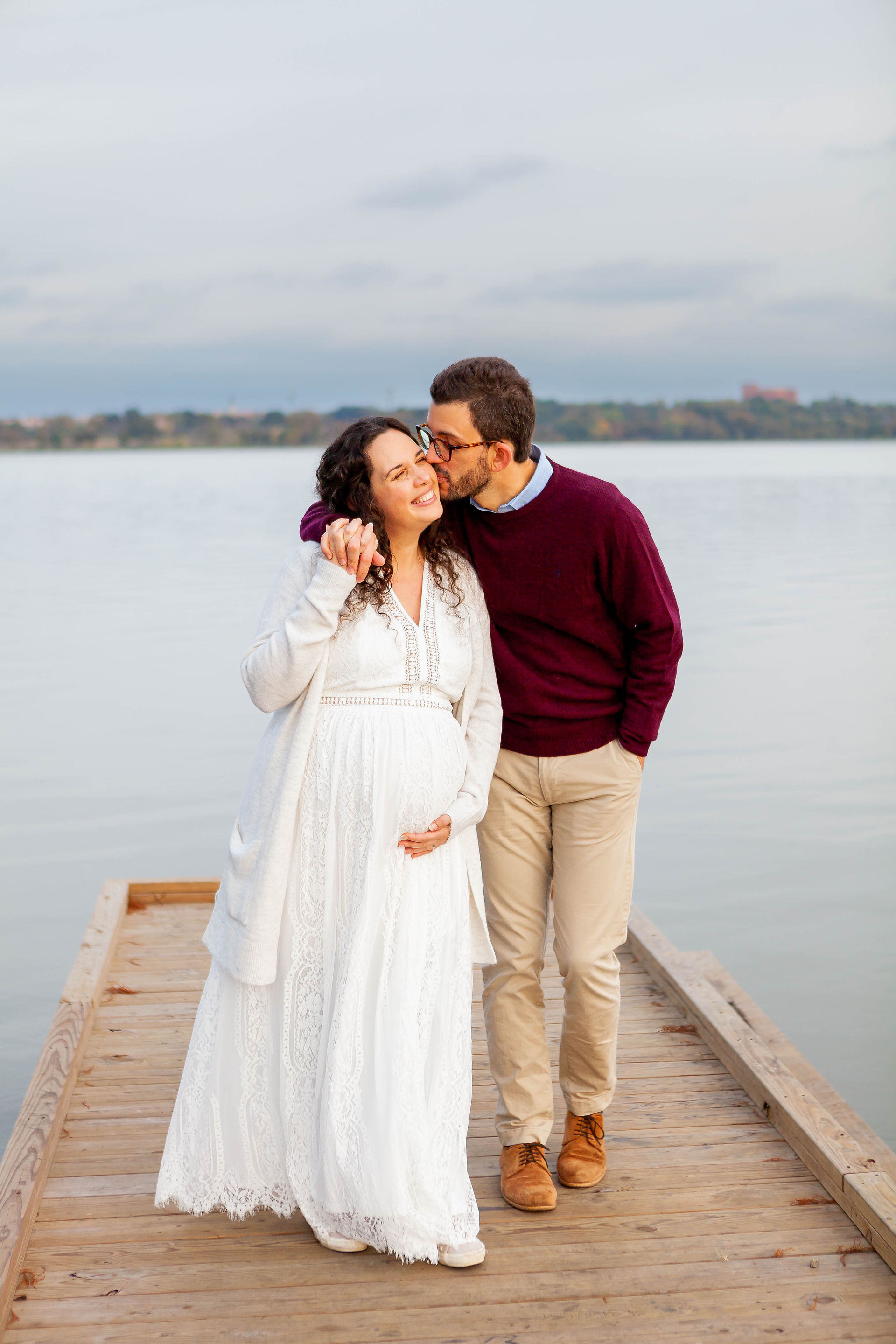 A lovely couple walks along a short pier towards the camera while holding hands. The husband is kissing his smiling wife on the cheek as she caresses her growing baby bump.  Image by Photography of Sarah Mae.