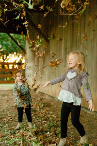 Two girls are playing in the fall leaves beside a rustic barn at the A.W. Perry Homestead. Having tossed the leaves into the air the older sister has her eyes closed and is smiling as the leaves rain down upon her face.